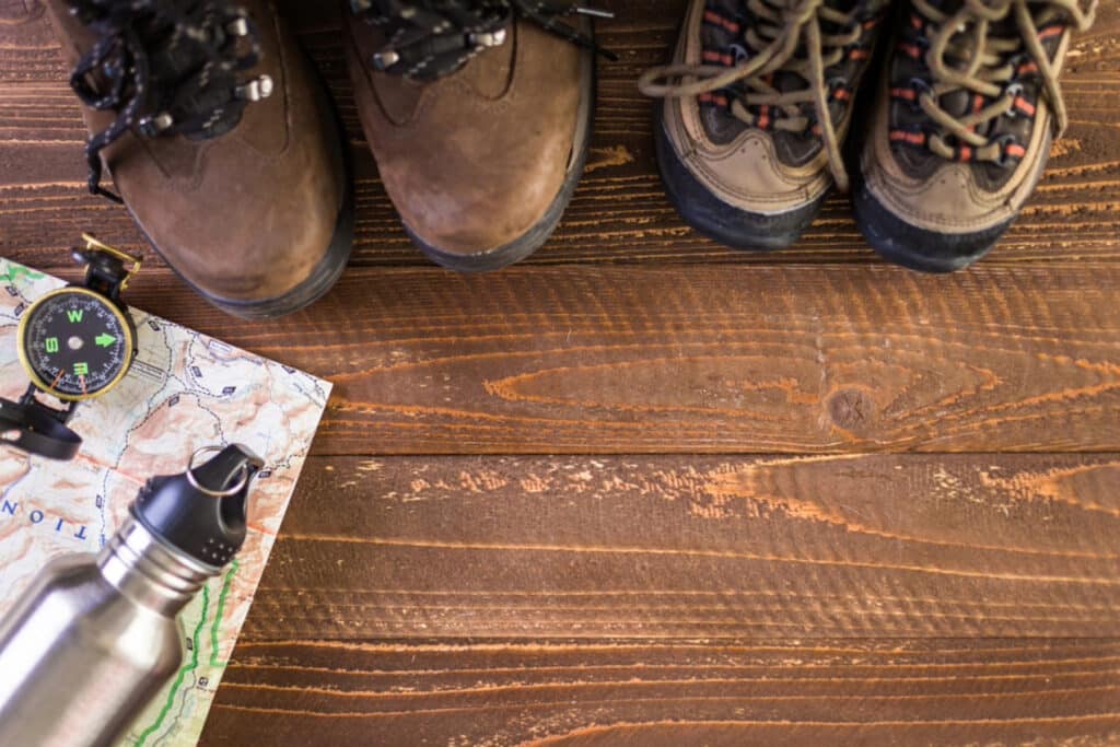 View of hiking shoes, a map, a compass, and water bottle on a wooden floor.