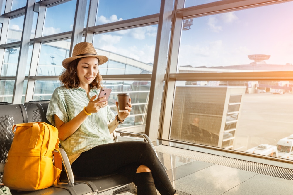 A woman sitting on an airport bench, focused on her phone, surrounded by travelers and luggage.