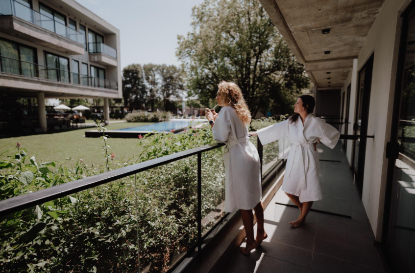 Two women in robes stand on a balcony, overlooking a serene pool below.