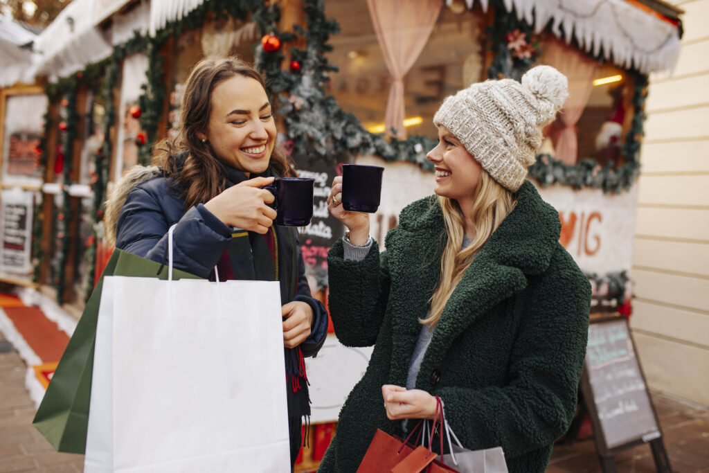 Two women enjoy coffee while holding shopping bags, smiling and chatting in a lively outdoor setting.