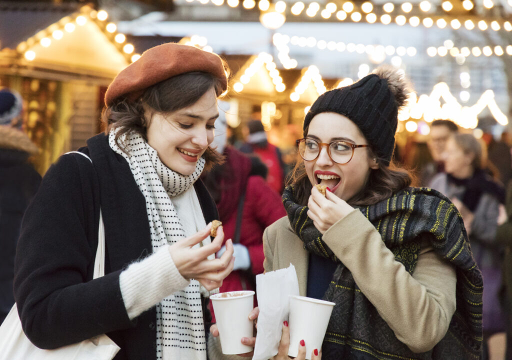 Two women enjoying food together at a festive Christmas market, surrounded by holiday decorations and cheerful atmosphere.
