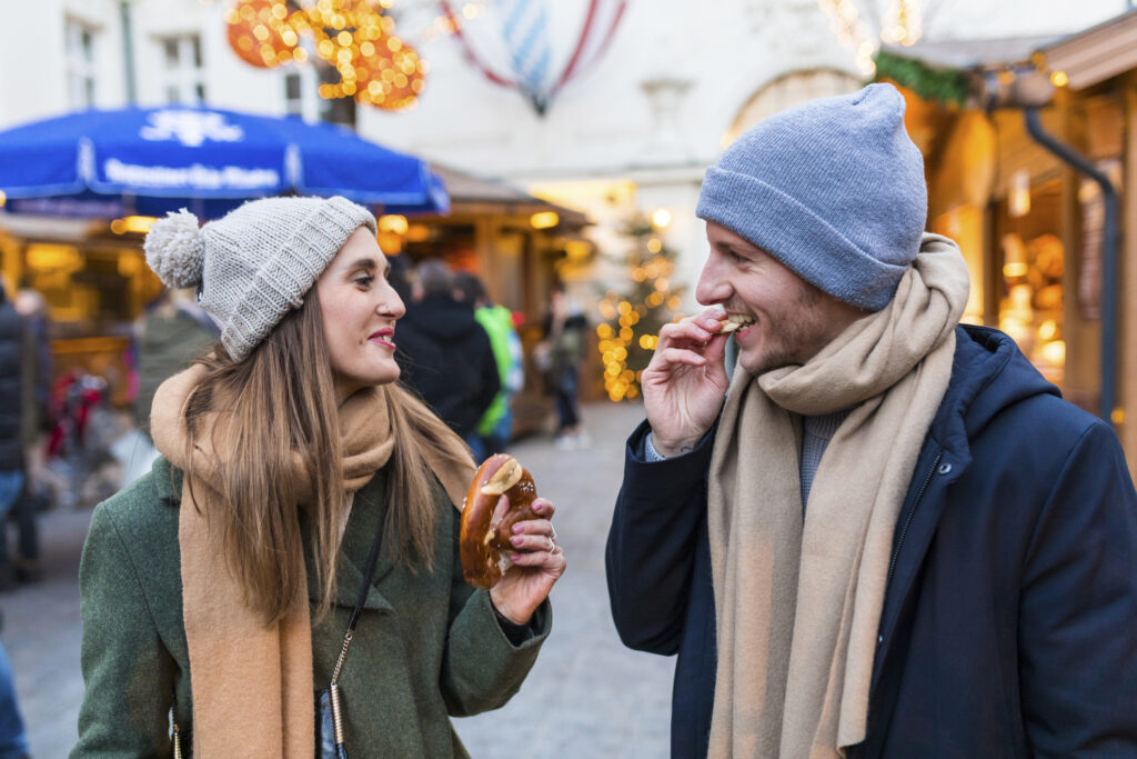 A couple enjoying a hotdog together at a festive Christmas market, surrounded by holiday decorations and lights.