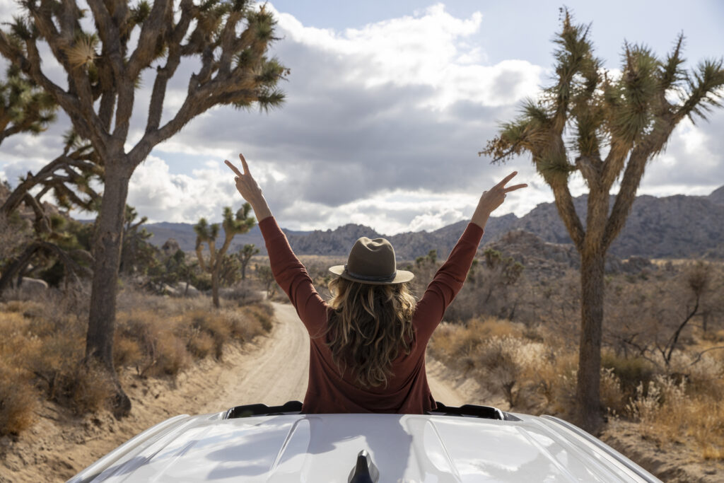 Woman wearing a hat, standing with arms raised in a peace sign pose, looking out of a car sunroof toward a desert landscape.