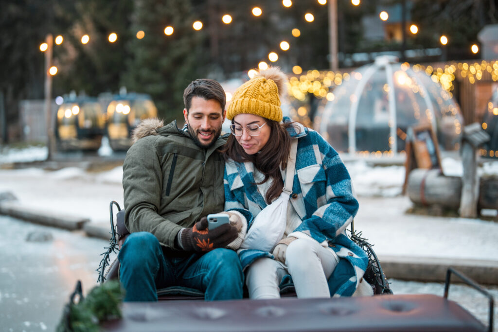 Candid portrait of young couple in winter attire sharing smartphone screen outdoors. Mid-aged man in green parka and woman in plaid coat and yellow beanie beneath string lights and dining domes.