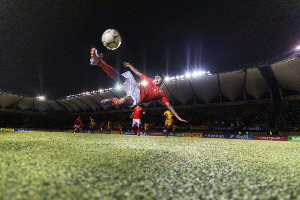 Wide shot low angle still of a young adult professional football player skillfully jumping up and kicking a soccer ball bicycle kick style in mid-air while illuminated by stadium floodlights during an international football match