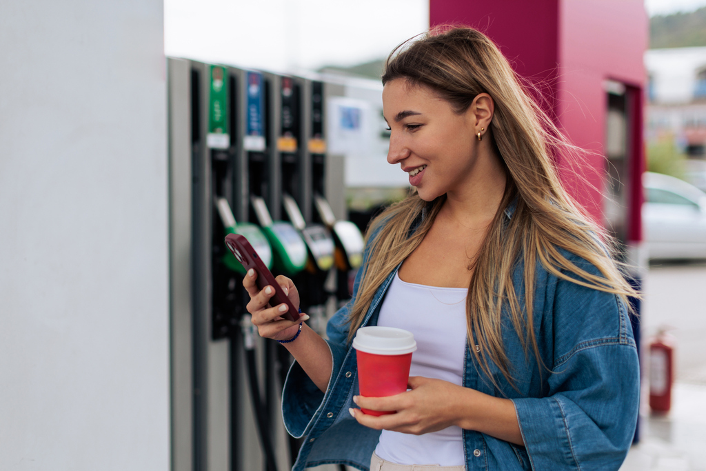 A woman stands at a gas station, sipping coffee and checking her phone.