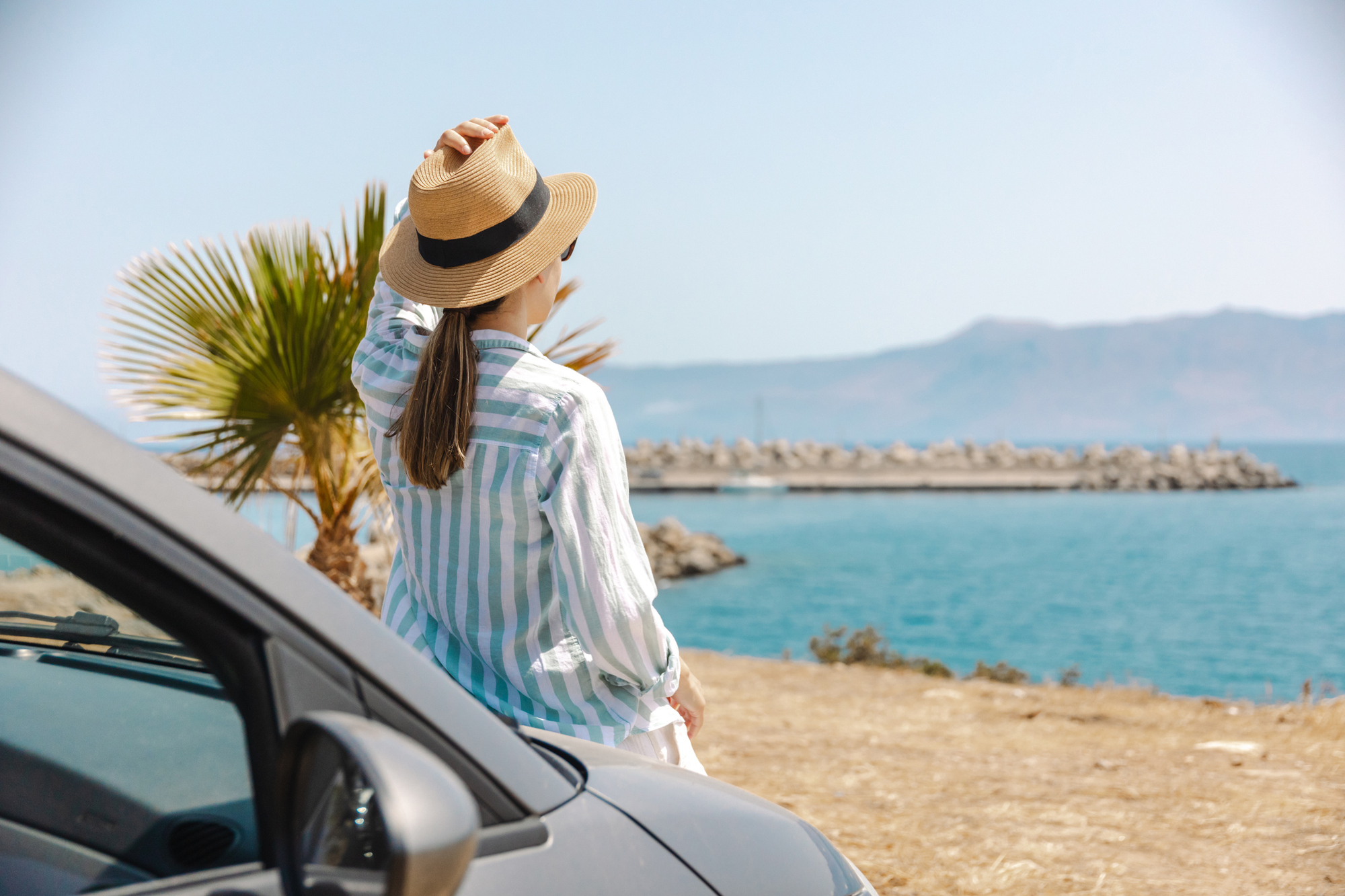 A woman wearing a hat stands beside a car, with the ocean visible in the background.