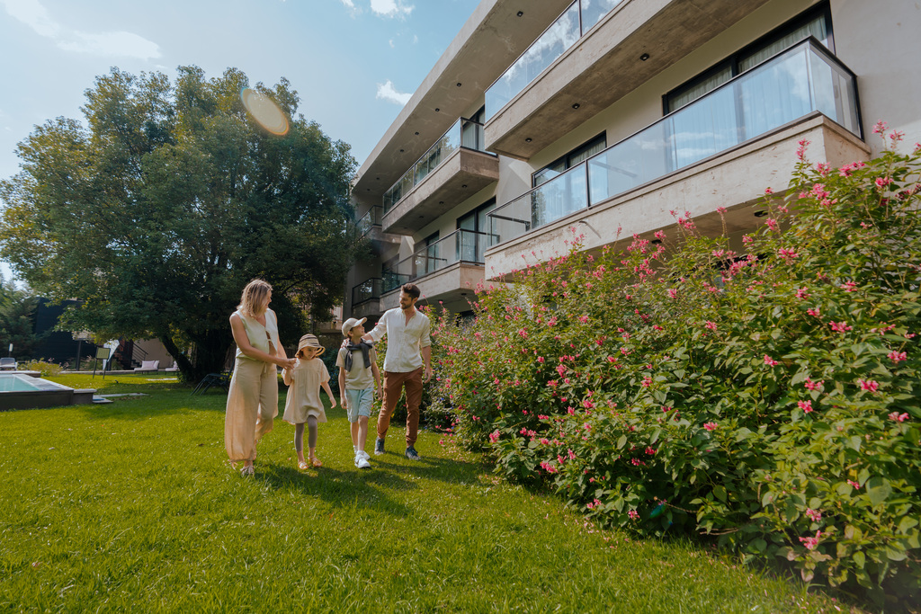 A family of four walks together on green grass near a modern building, enjoying a sunny day outdoors.
