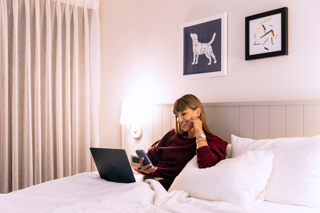 A woman sitting on a bed, focused on her laptop, with a cozy bedroom setting in the background.