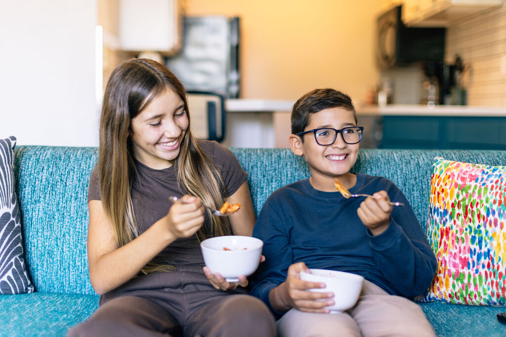 Two children on a couch, each with a bowl of cereal, smiling as they eat.