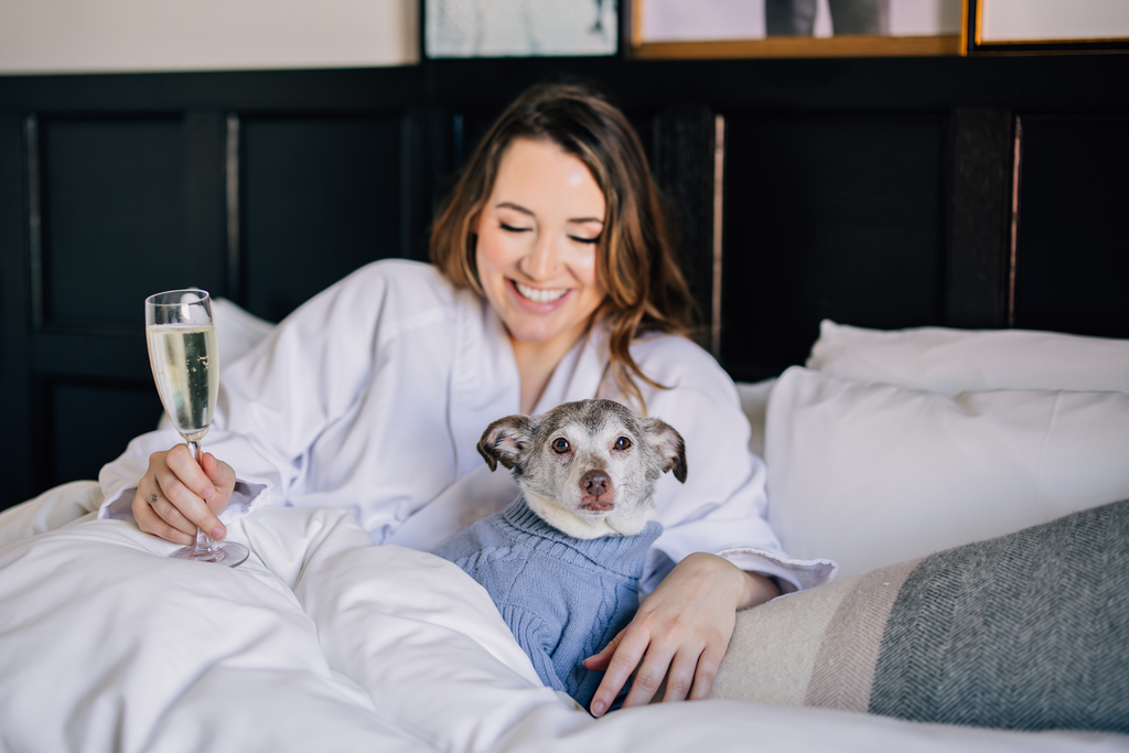 A woman relaxing in bed with a dog beside her, holding a glass of champagne, enjoying a cozy moment.