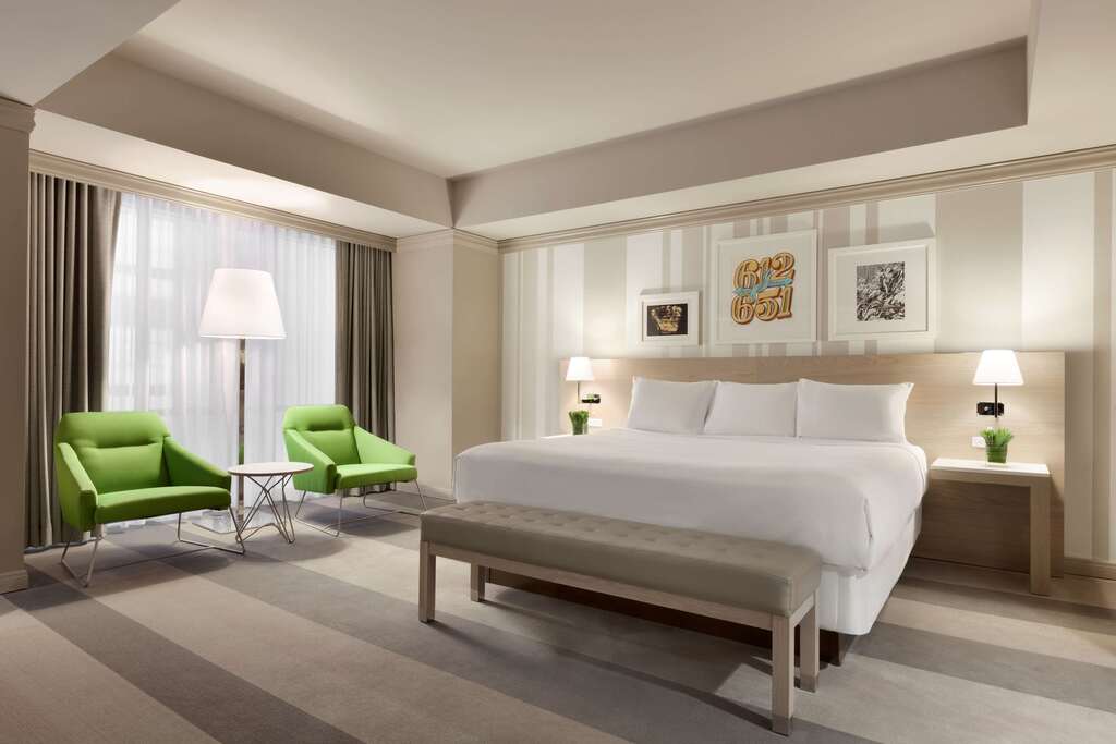 A hotel room featuring a bed, a chair, and two green chairs, creating a cozy and inviting atmosphere.
