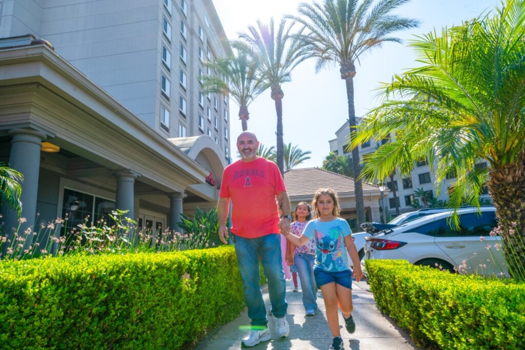 A man and two children stroll along a sidewalk in front of a hotel, enjoying a sunny day together.