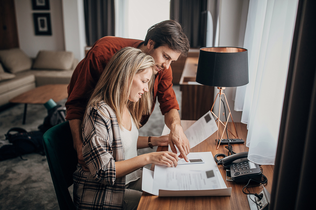 A man and woman are seated at a desk in a hotel room, engaged in reviewing and organizing paperwork.