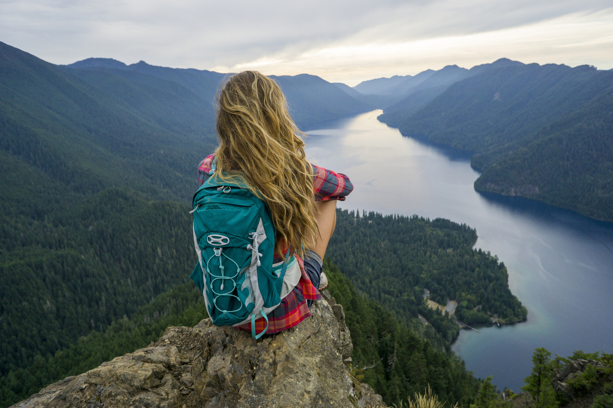A woman with a backpack is seated on a rock, overlooking a calm lake framed by lush greenery and distant hills.