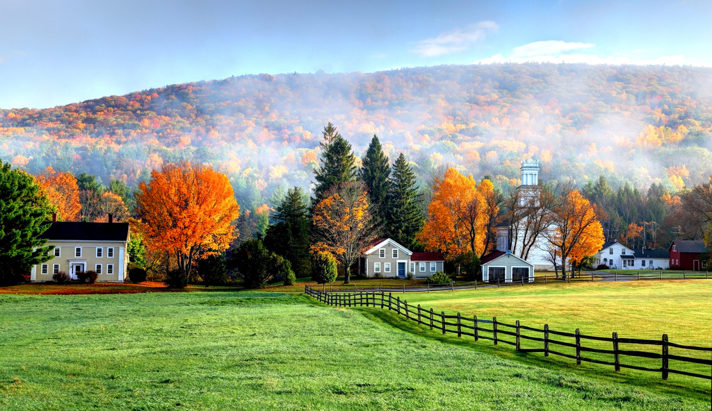 A scenic field bordered by a wooden fence, with a farm and trees in the background under a clear blue sky.