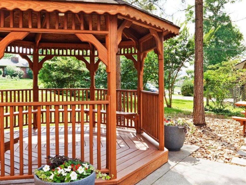 A gazebo featuring a picnic table surrounded by benches, set in a green outdoor space.