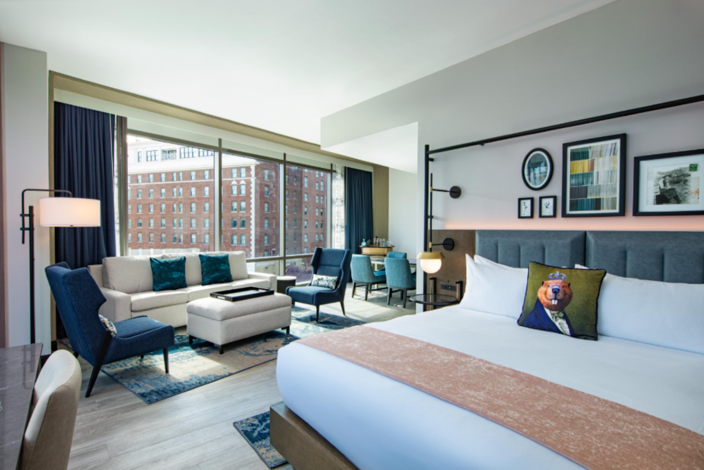 A hotel room featuring a bed, desk, chair, and a large window showcasing a city view.