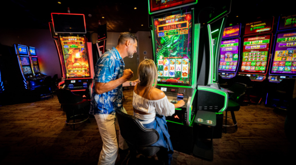 A man and woman are seated at slot machines in a vibrant casino, focused on their gameplay.