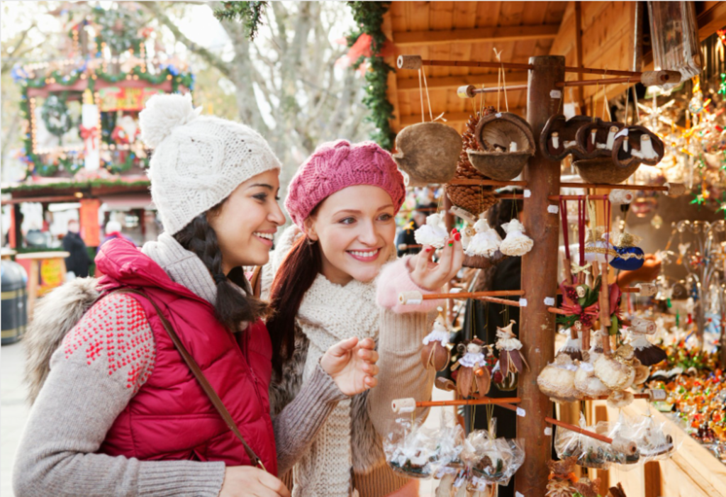 Two women admire Christmas decorations at a festive market, surrounded by colorful lights and holiday displays.