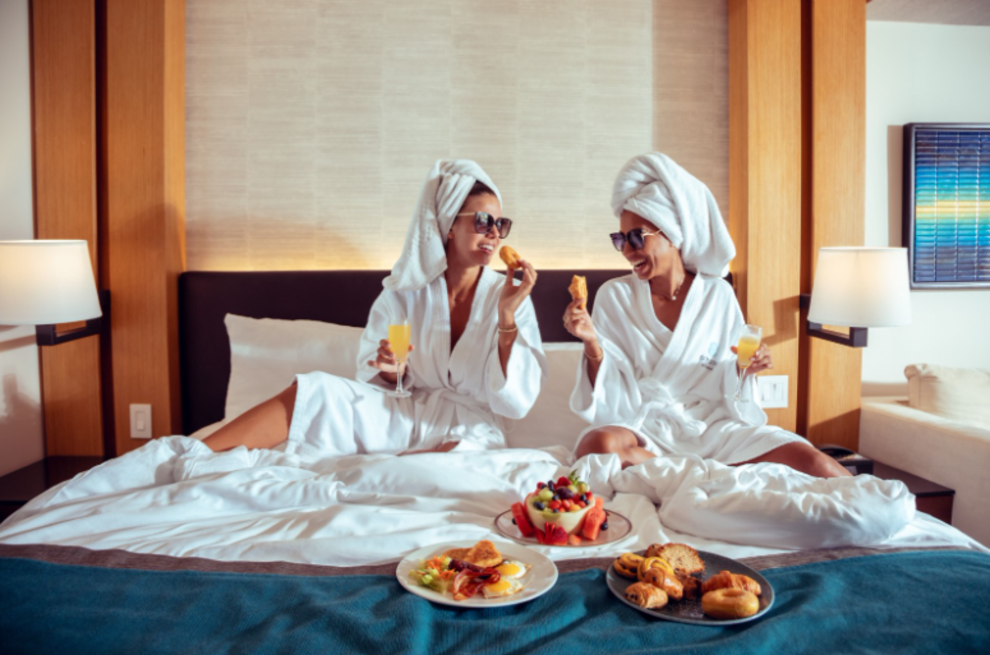 Two women in bathrobes enjoy breakfast together on a bed, surrounded by pillows and a cozy atmosphere.