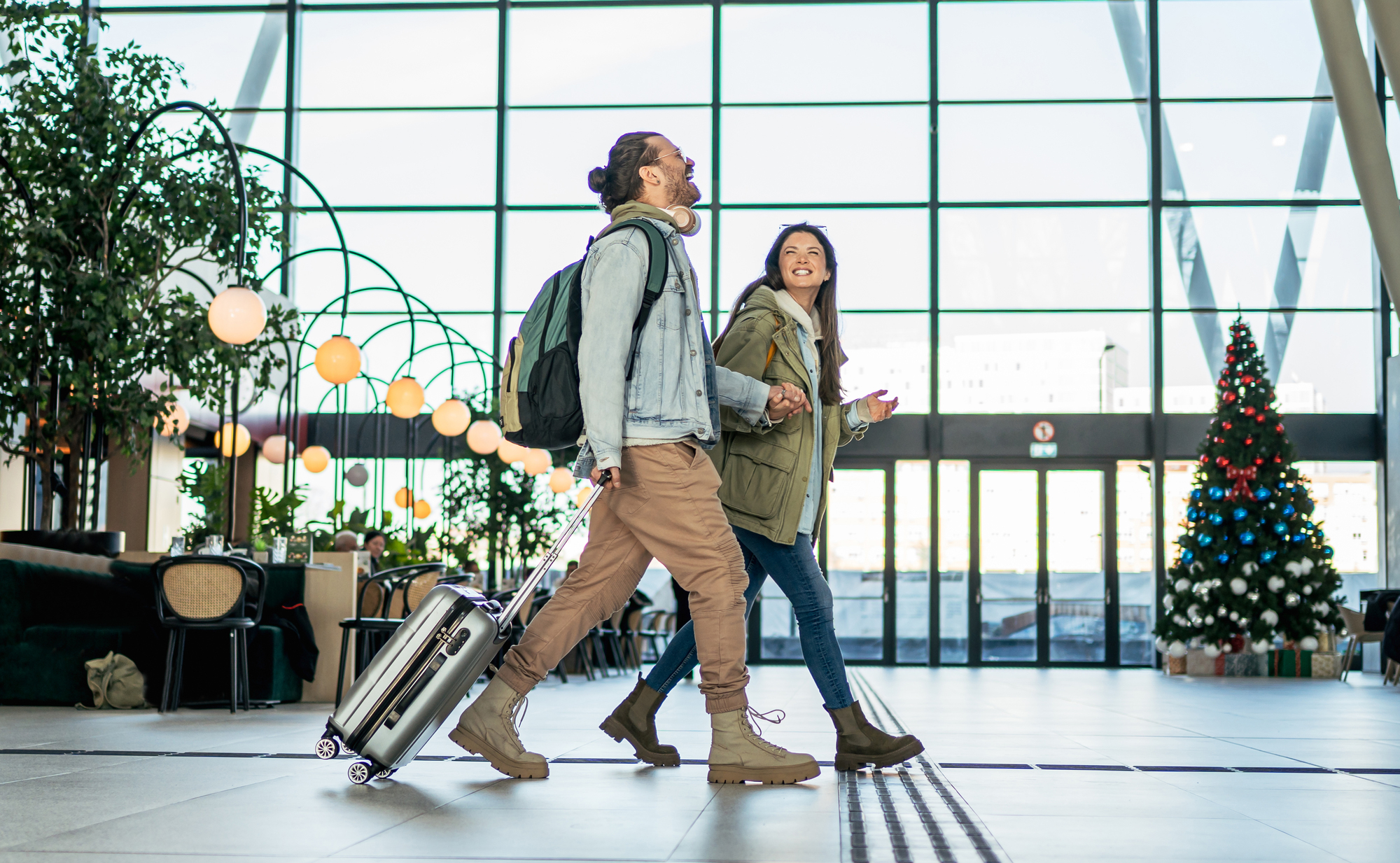 A couple walks through an airport terminal, each carrying luggage and looking at their surroundings.