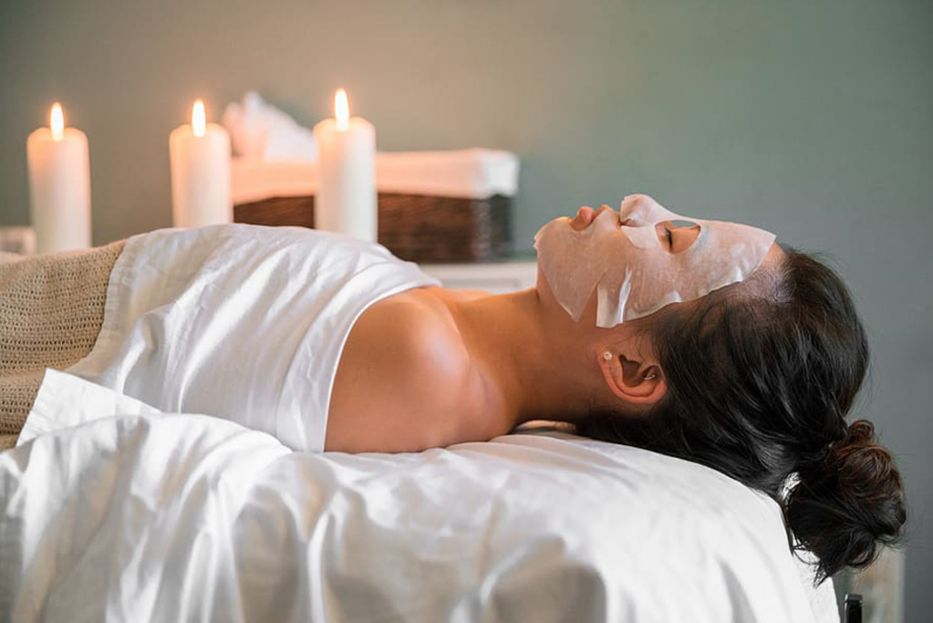 A woman relaxes in a spa room, wearing a facial mask and surrounded by calming decor.