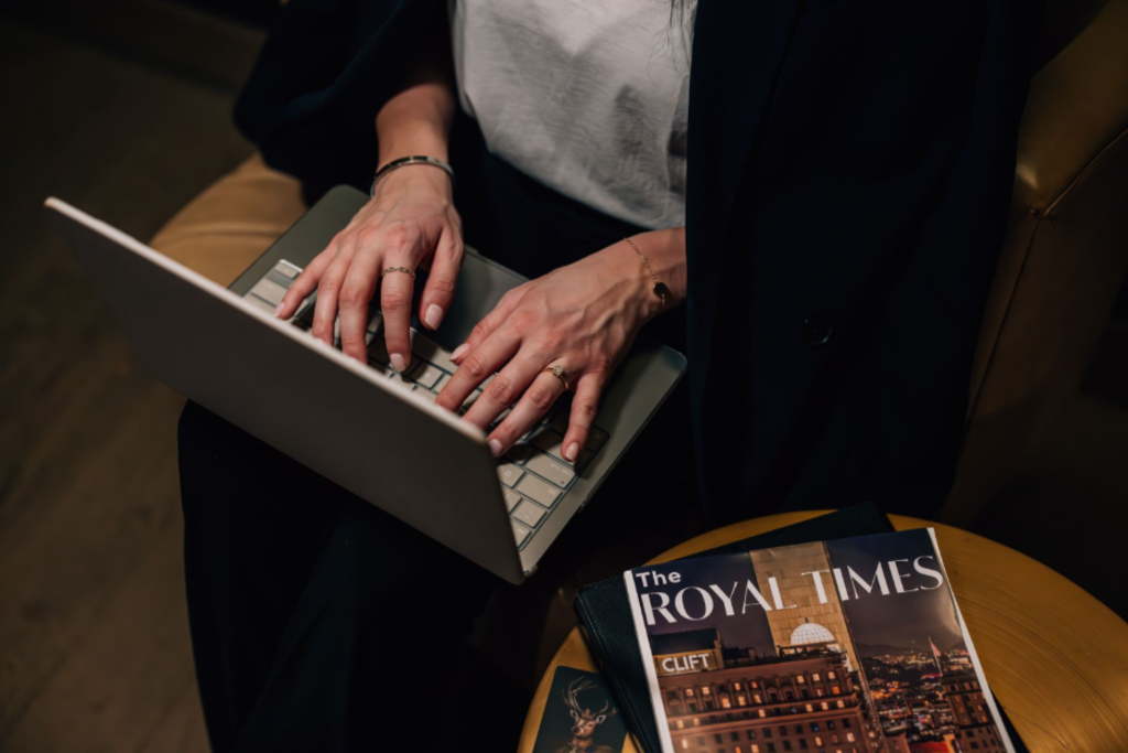 A woman sitting on a chair, focused on her laptop, with a cozy indoor setting in the background.