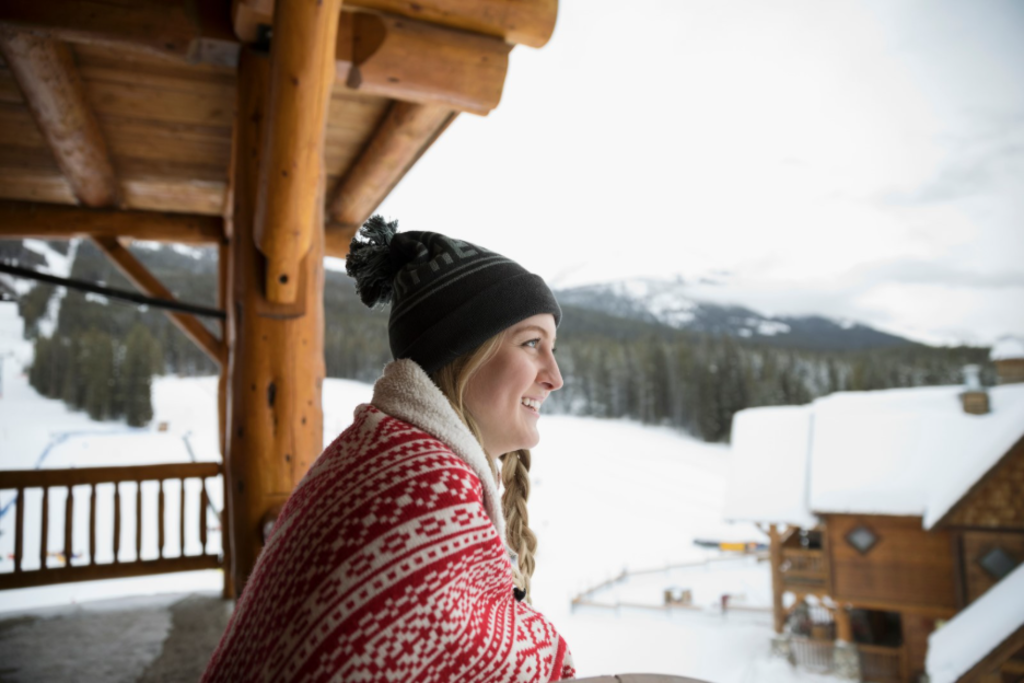 A woman relaxes on a porch, overlooking a ski lodge surrounded by snow-covered mountains.